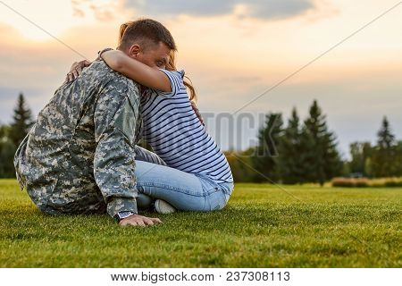 Soldier Hugging With Wife Against Evening Sky Background. Woman Ebracing His Husband Sitting On The 