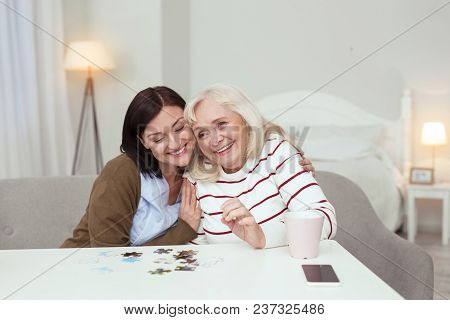 Interesting Puzzle. Cheerful Elder Woman And Caregiver Hugging While Gathering Puzzle