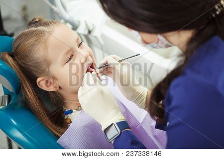 Child Lies In Comfortable Dentis Chair And Goes Through Procedure While Female Doctor In Mask And Ru