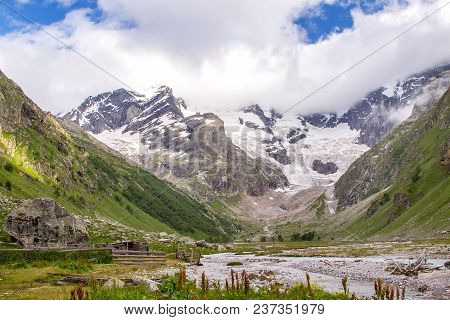 Colorful View Of Green And Snowy Mountains With Elbrus Summit Under Fluffy White Clouds.