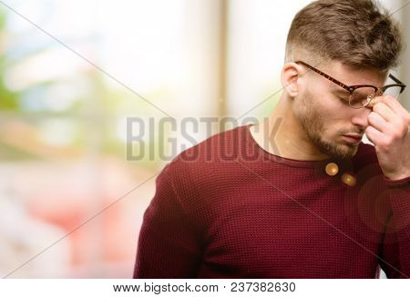 Handsome young man with sleepy expression, being overworked and tired, rubbes nose because of weariness