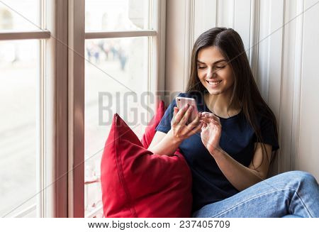 Sexy Brunette Girl Wears Blue T-shirt Holds Smartphone Sitting On Wide Windowsill With Big Red Pillo