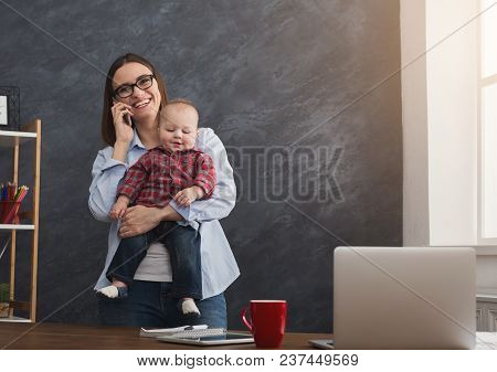 Happy Mother Working At Office With Her Baby. Young Woman Talking On Phone And Using Laptop While Sp