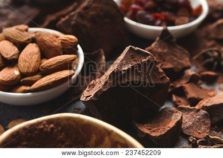 Dark Chocolate Bar Broken In Pieces On Dark Background, Cocoa Powder, Various Nuts And Raisins In Bo