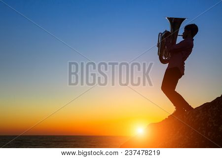 Silhouette of musician with the tuba/trumpet on rocky sea coast during amazing sunset. 