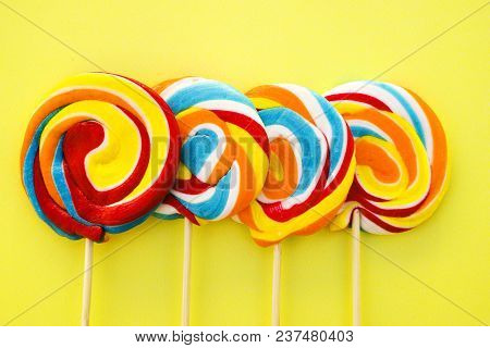 Colorful Hard Candy Lollipop On Yellow Pastel Background. Rainbow Sweet Pattern On Stick Flying Arou