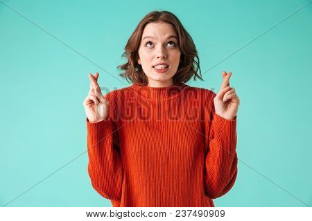 Portrait of a worried young woman dressed in sweater holding fingers crossed for good luck isolated over blue background
