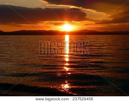 A Beautiful Sunset Over The Lake. Red Sky, Sun, Clouds And Waves. The Mountains On The Horizon In Wh