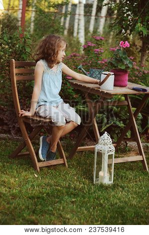 Happy Kid Girl Playing In Summer Garden, Holding Heranium Flower In Pot. Teaching Kids To Love Natur