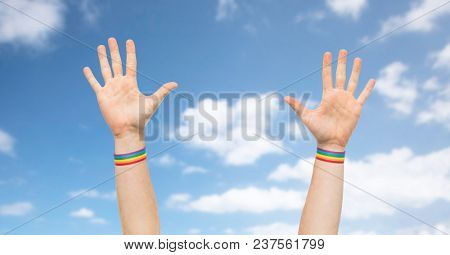 lgbt, same-sex relationships and homosexual concept - close up of male hands wearing gay pride awareness wristbands over blue sky and clouds background