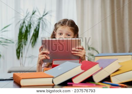 Little Girl Looking At The Tablet Pc In A Classroom. Colorful Books At The Table.