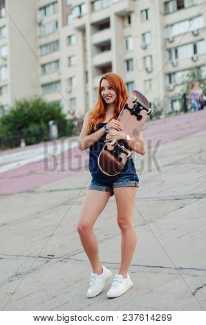Redhead Slim Girl In Jeans Shorts And Black T Shirt Posing With Skateboard In Urban Sciene.
