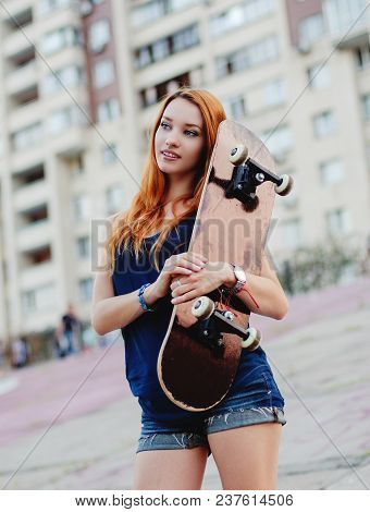 Redhead Slim Girl In Jeans Shorts And Black T Shirt Posing With Skateboard In Urban Sciene.