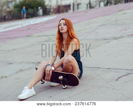 Redhead Slim Girl In Jeans Shorts And Black T Shirt Posing With Skateboard In Urban Sciene.