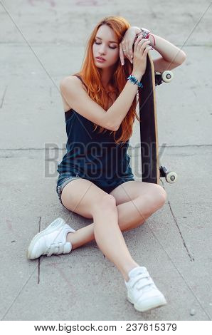 Redhead Slim Girl In Jeans Shorts And Black T Shirt Posing With Skateboard In Urban Sciene.