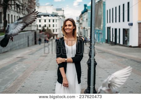 Young Woman Stands At A Street. Pigeons Flyes Around The Girl. Beautiful Girl Walks Along The Street