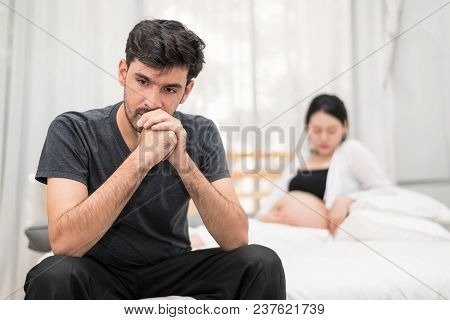 Close Up Of Serious Man Sitting On Bed With Blur Of Unhappy Pregnant Woman With Big Belly.needless P