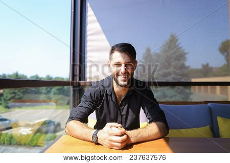 Male Manager Sitting At Table On Sofa And Smiling, Wearing Black Shirt. Concept Of Business Occupati
