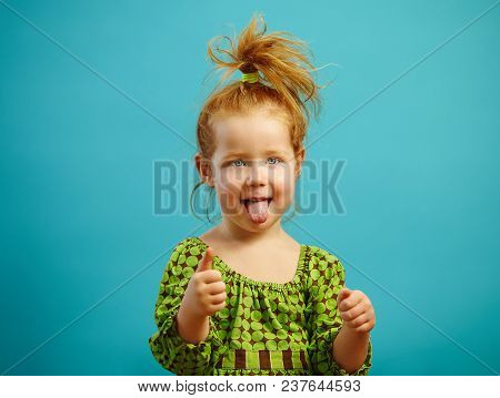 Portrait Of Three Year Old Girl Stuck Her Tongue Out And Shows Thumb Up, Has Red Hair, Dressed In Gr