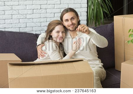 Portrait Of Happy Couple Holding Keys Embracing Sitting On Couch With Boxes In New Home, Smiling Ren