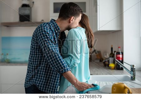 Man Kisses A Woman In The Neck In The Kitchen Interior. Co-wash Dishes, Newlyweds In A New Home