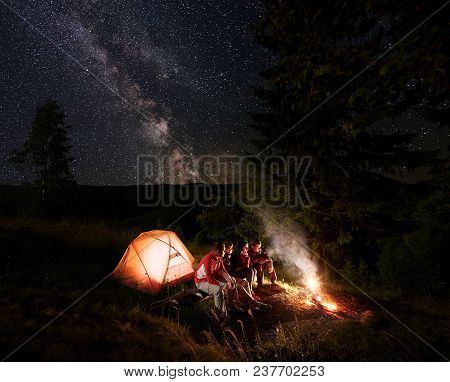 Four Friends Hikers Having A Rest On A Log Near Illuminated Orange Tent, Enjoying The Flame Of A Cam