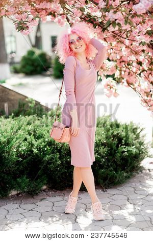Beautiful Young Woman With Curly Pink Hair And Sunglasses Near The Blossoming Spring Tree