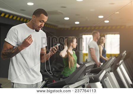 Young African-american Man With Smartphone Running On Treadmill In Gym. Fitness Guy Jogging In Fitne