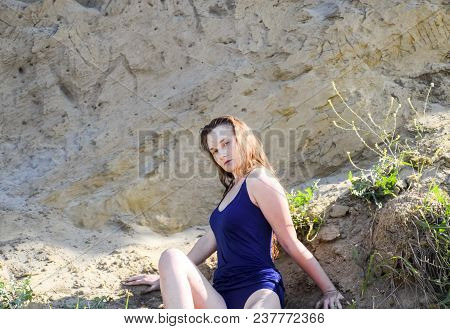 Beautiful Blonde In A Blue Bathing Suit Sits On A Steep Bank. Coast Of Clay And Sand.