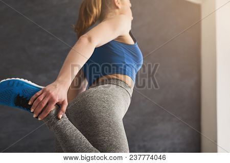 Young Woman Warming Up Before Training At Gym. Unrecognizable Girl Stretching Her Leg At Gray Backgr