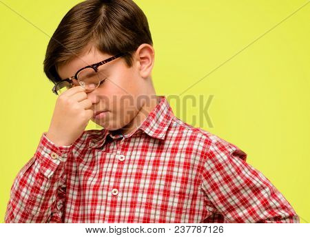 Handsome toddler child with green eyes with sleepy expression, being overworked and tired, rubbes nose because of weariness over yellow background