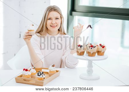 Beautiful Young Woman In Pink Sweatshirt Having Cupcakes While Sitting Near Window At Cookery