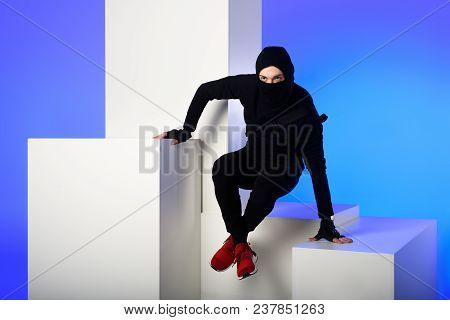Ninja In Black Clothing With Katana Behind Getting Across On White Block Isolated On Blue