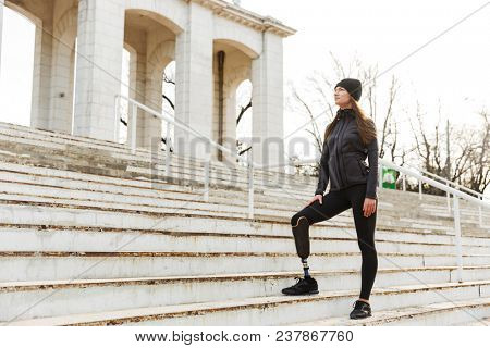 Photo of atlethic disabled woman in sportswear with prosthetic leg standing at the stairs and looking aside outdoor