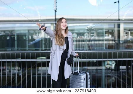 Woman Stopping Taxi With Raised Hand, Valise And Smartpone Near Airport. Concept Of Business Trip An