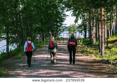 Three Women With Backpacks And Walking Sticks Make A Promenade Along The Sandy Path Along The Shore 