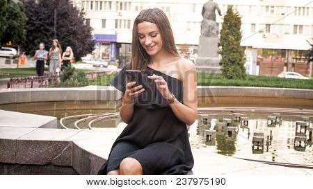 Portrait Of Smiling Woman Sitting At Fountaing And Using Mobile Phone
