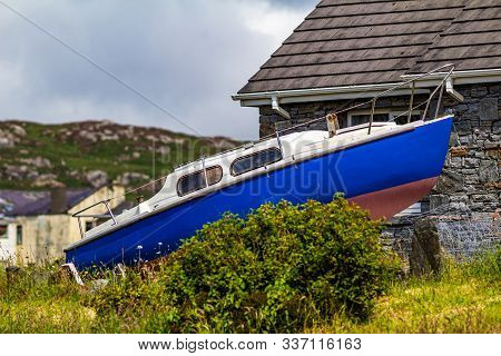 Small Boat Next To A House Near The Pier Of The Port Of Clifden With A Mountain In The Blurred Backg