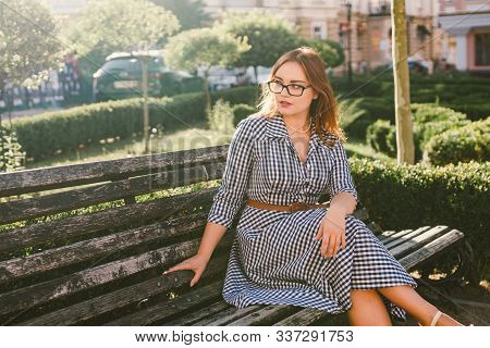 Young Woman On The Street In Summer Dress Sitting In Summer Park On Bench. Beautiful Young Woman In 