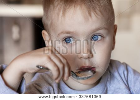 Cute Caucasian Baby Boy With Blue Eyes And Blond Hairs Eating Porridge. Serious Thoughtful Look, Chi