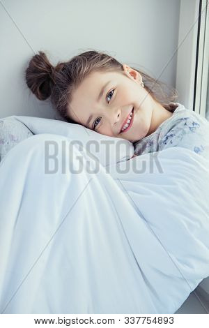 A little girl sittiing on a windowsill on a pillow covered with a blanket. Childhood. Kid's fashion.