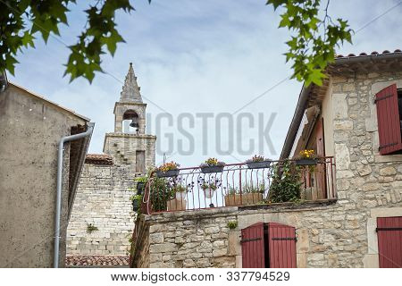 Barjac, France. Bell Tower, Building Facade With Wooden Shutters And A Terrace With Flowers. View Of