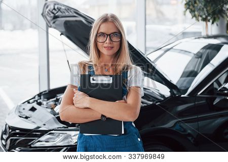 Our Mehanics Works On Your Vehicle. Manager With Notepad, In The White Shirt And Blue Uniform Stands
