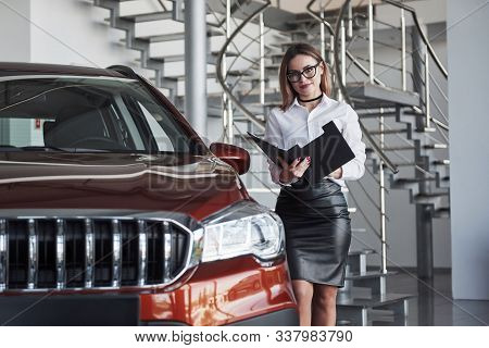 Woman In Eyewear, Black Skirt And White Shirt Stands Near The Red Automobile.