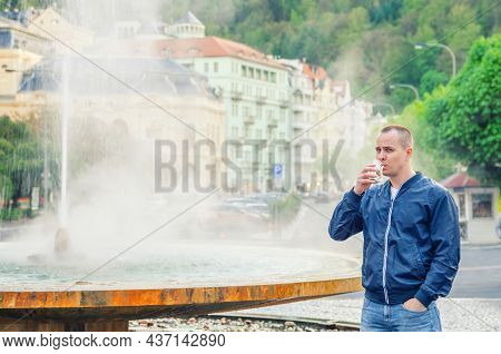 Young Man Tourist With Blue Jacket Look At Camera And Drink Therapeutic Mineral Water From Cup, Foun