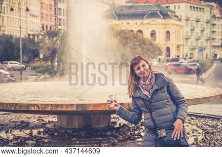 Young Woman Tourist With Grey Jacket Posing And Hold Therapeutic Mineral Water In Cup Near Fountain 