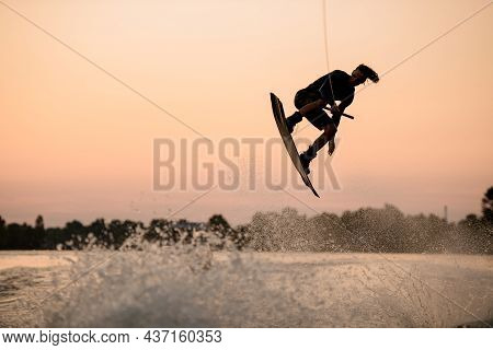 Active Man Jumping Over Splashing Wave On Wakeboard Holding On To The Rope. Water Sports Activity.