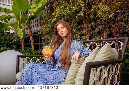 Stylish Woman With Glass Of Non-alcoholic Cocktail Sitting On Couch In Rooftop Garden Of Luxury Hote