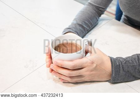 Woman's Hands In Grey Long Sleeve Shirt Holding Cup Of Hot Chocolate On The White Marble Countertop 