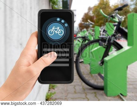 technology, transport and sustainability concept - close up of hand holding smartphone with mobile app and bicycle icon on screen over electric bike parking and charging station in city on background